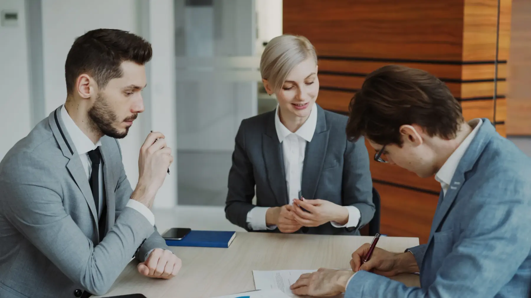 Três pessoas conversando em volta de uma mesa de reunião, uma delas assinando um papel