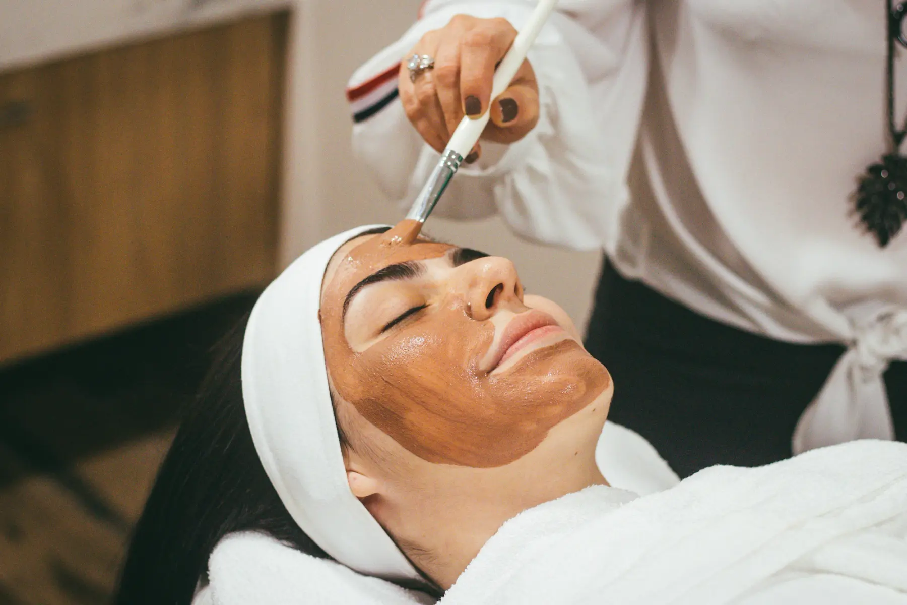 Esteticista aplica com pincel uma máscara de argila no rosto de uma cliente de olhos fechados e faixa branca no cabelo