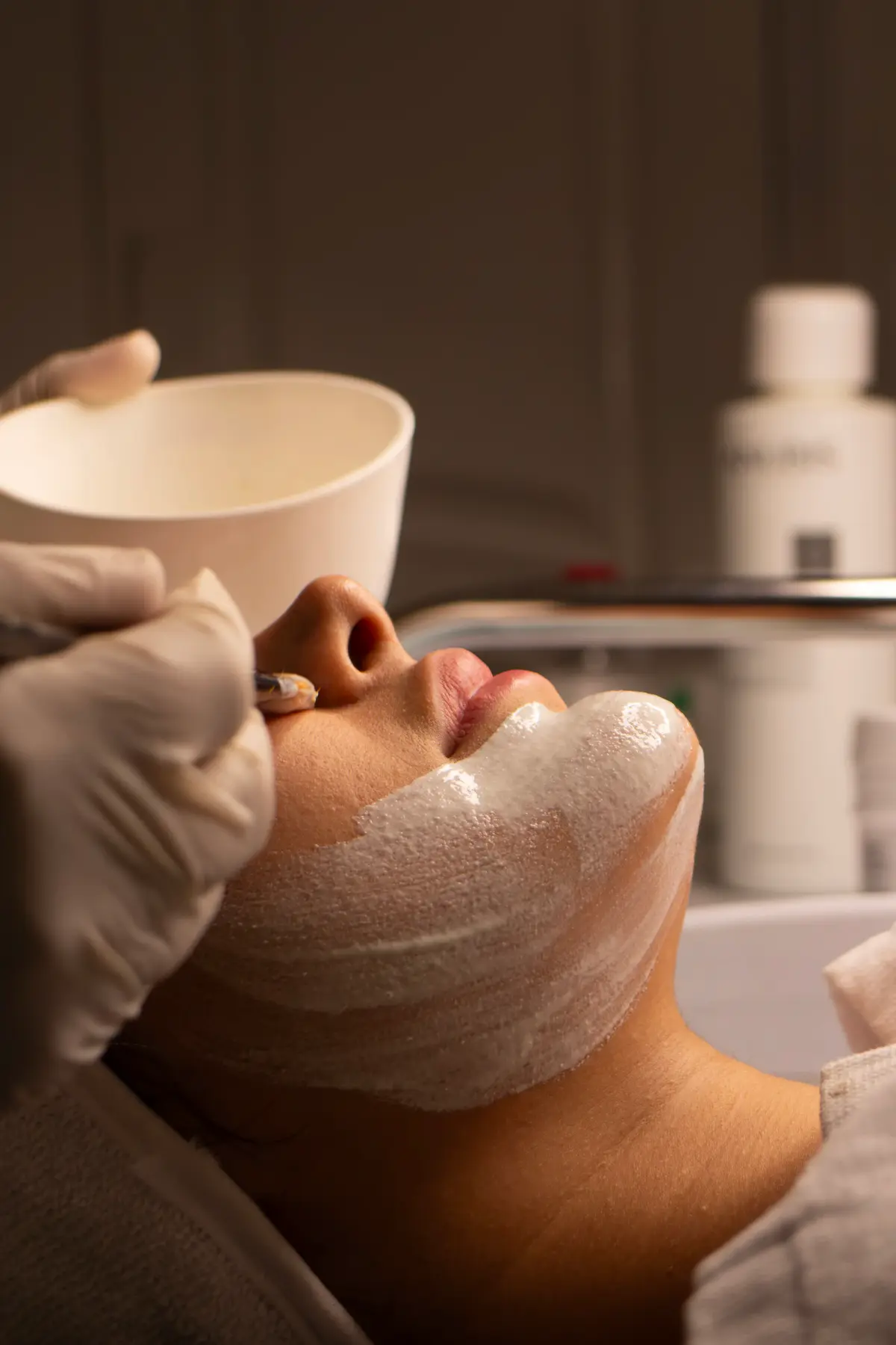 Esteticista de luvas aplica máscara branca no queixo de uma cliente deitada, com pincel e cumbuca