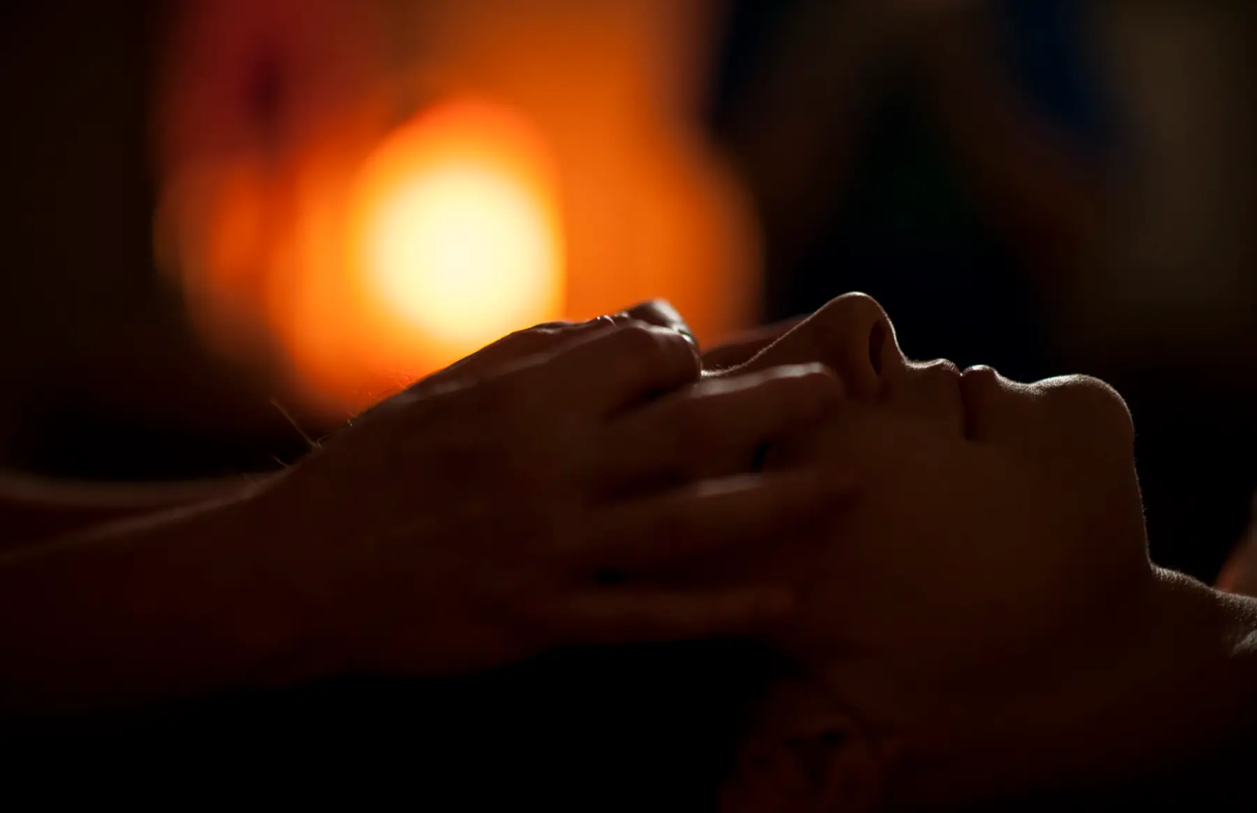 Mãos fazem massagem facial em uma mulher de olhos fechados, à luz de uma vela desfocada ao fundo