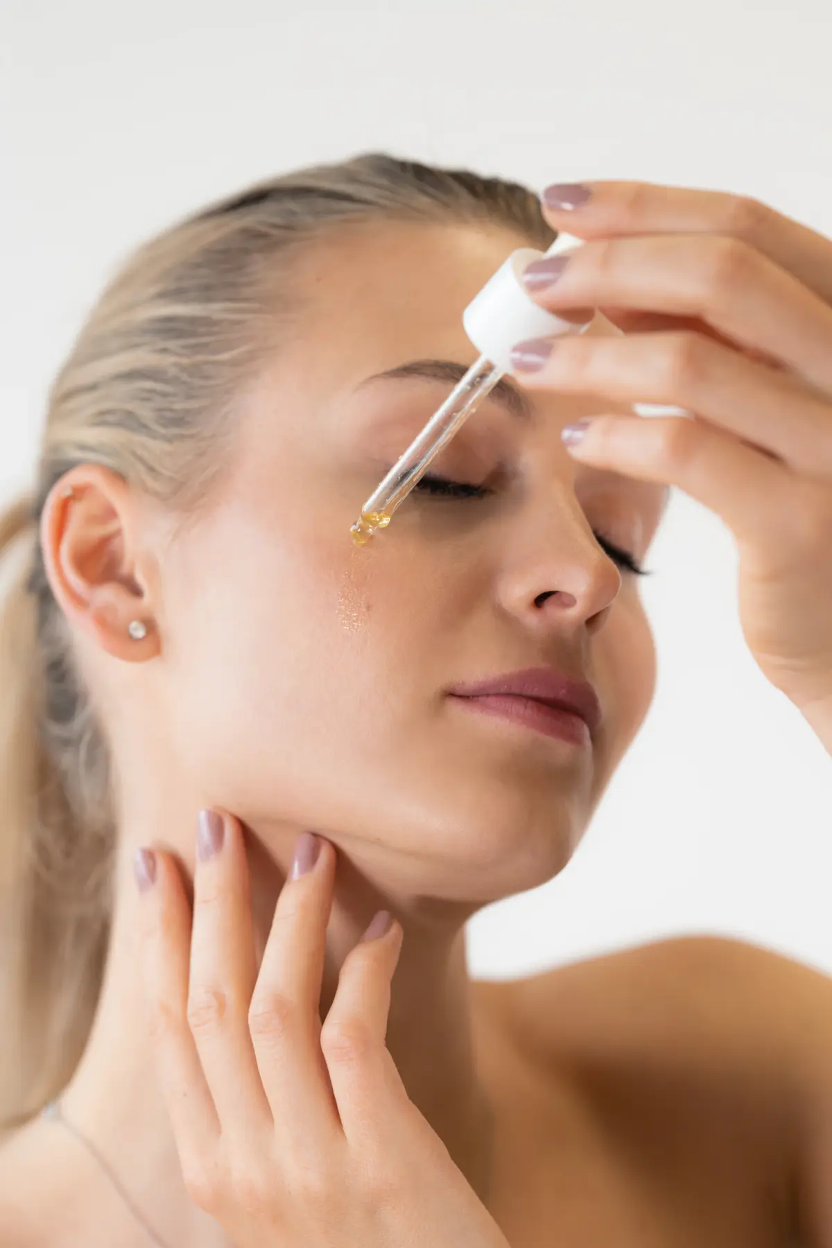 Mulher de olhos fechados pinga uma gota de sérum dourado no rosto com um conta-gotas