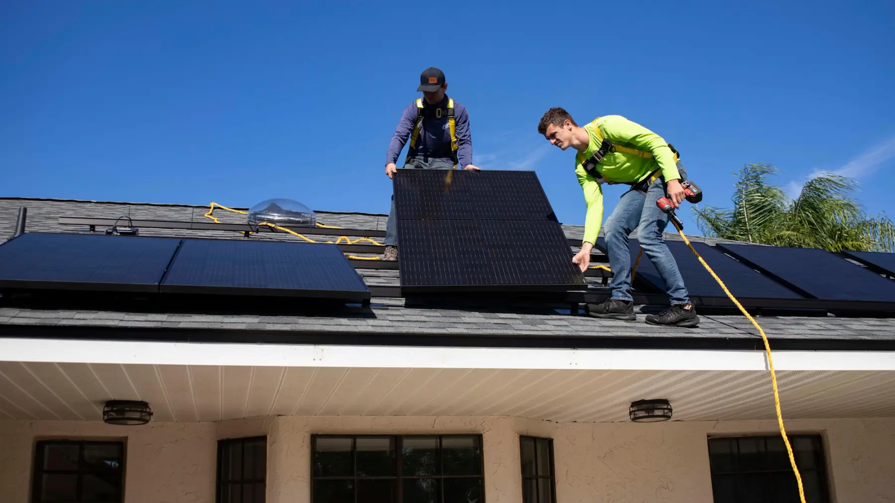 Dois instaladores com cinto de segurança posicionam uma placa solar no telhado de uma casa, sob céu azul