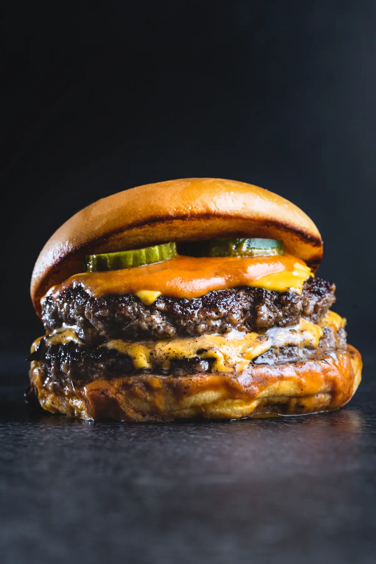 Smash burger duplo em close, com cheddar derretido e picles, sobre fundo escuro