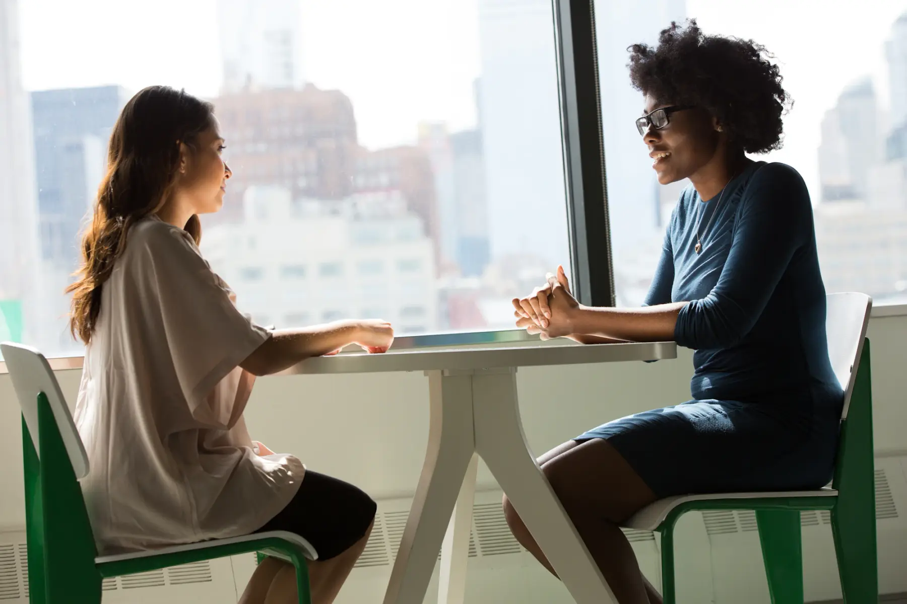 Duas mulheres conversando à mesa, perto de uma janela ampla com luz natural