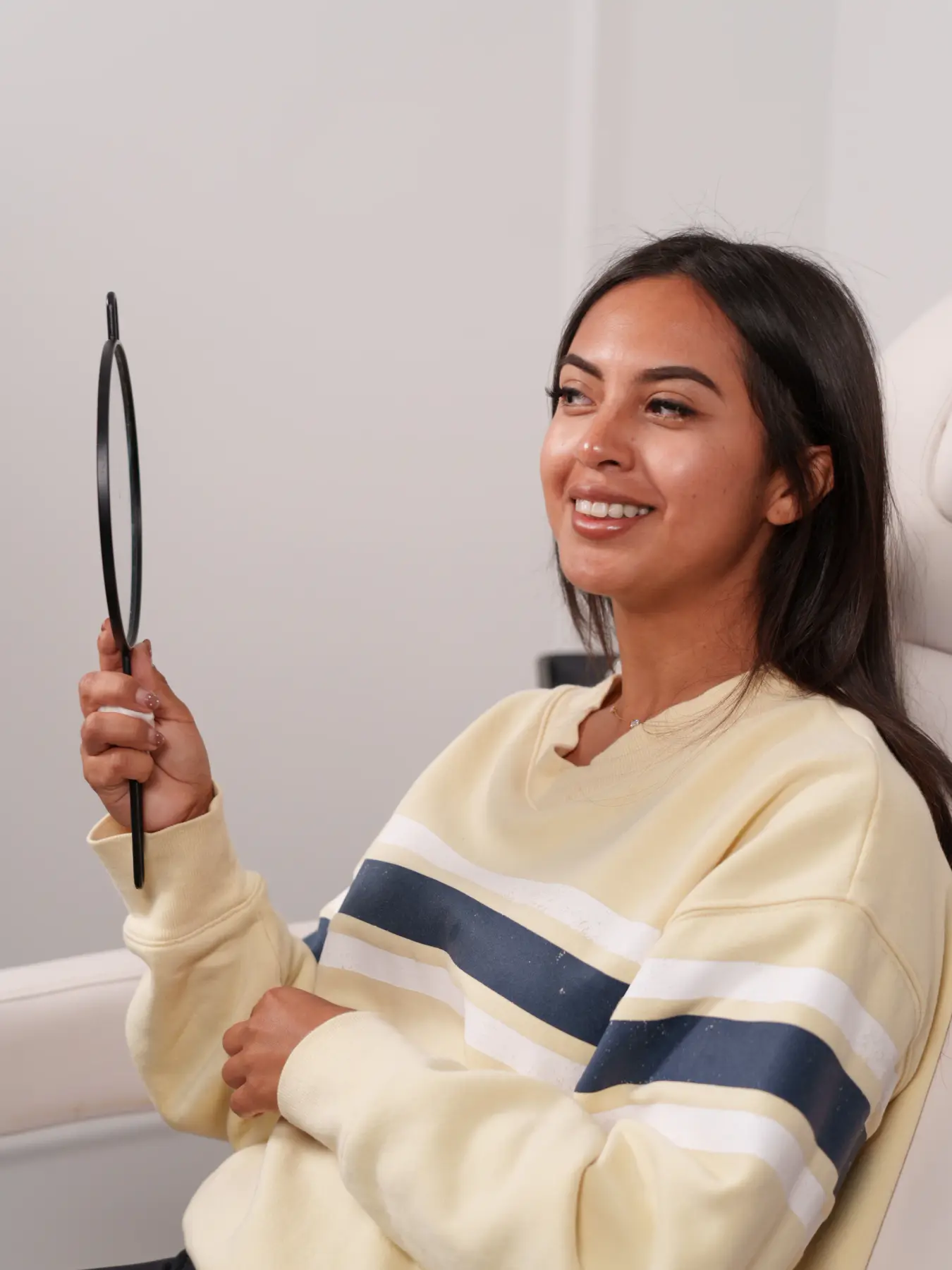 Paciente sorrindo enquanto se olha em um espelho de mão, sentada na cadeira do consultório