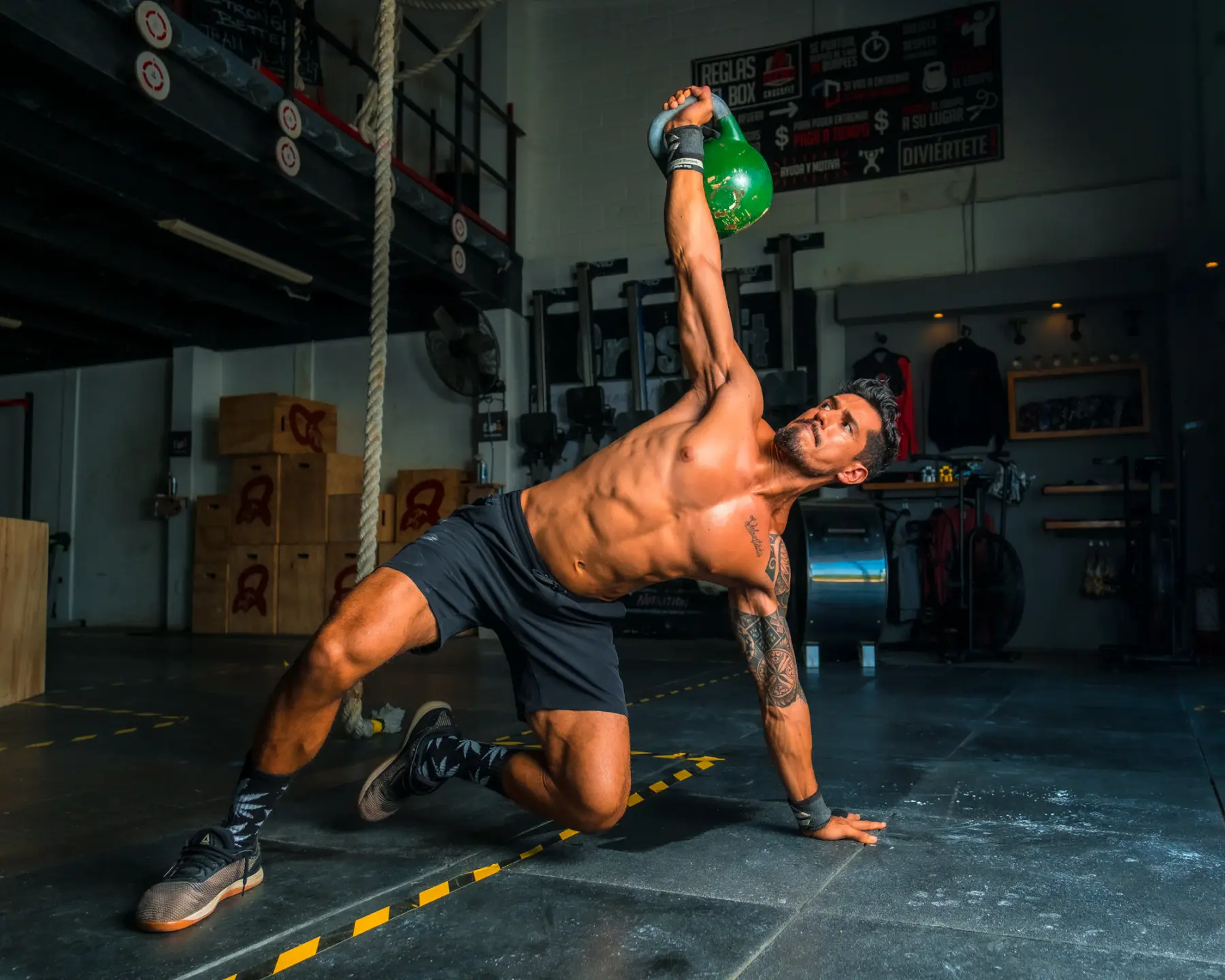 Homem girando o tronco com um kettlebell erguido acima da cabeça, apoiado em um joelho no chão do estúdio